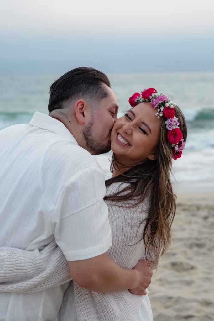 Guy kissing girl's cheek on a California beach. Girl is wearing a flower crown. 