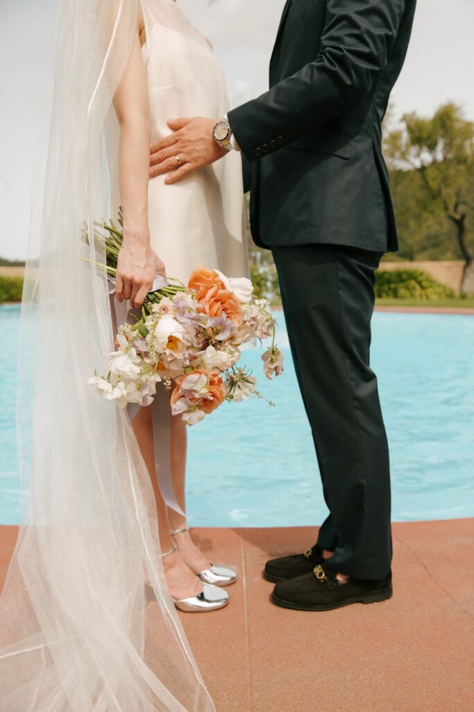 Bride and groom standing poolside, bride holding peach bridal bouquet with cathedral veil, California wedding photography