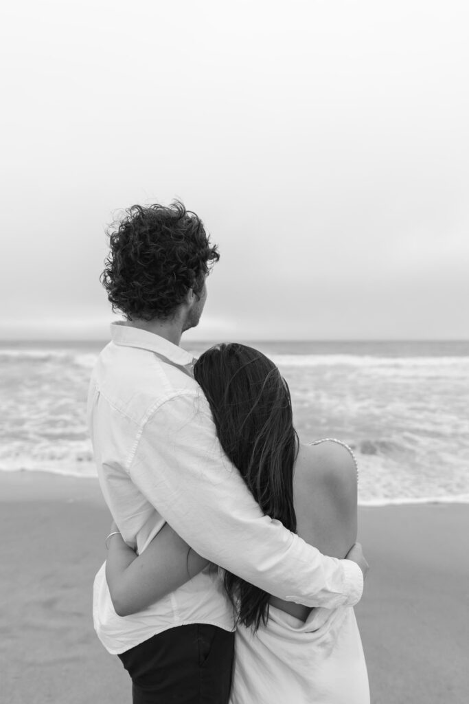 Couple embracing on Half Moon Bay beach, black and white engagement photography Bay Area