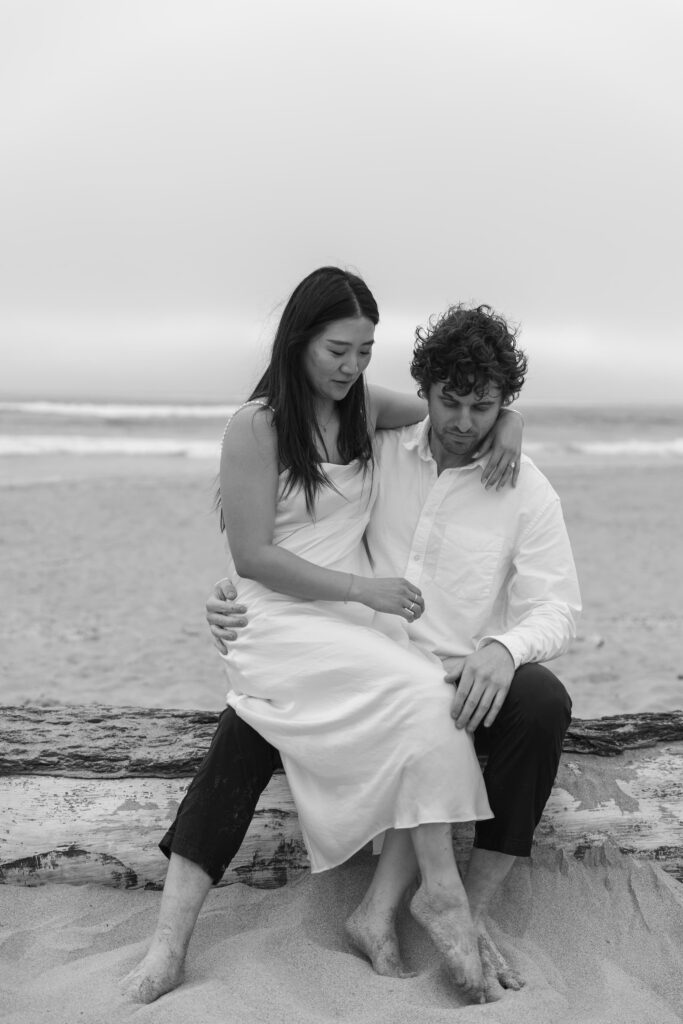 Couple sitting on driftwood at Half Moon Bay beach, black and white engagement photography Bay Area