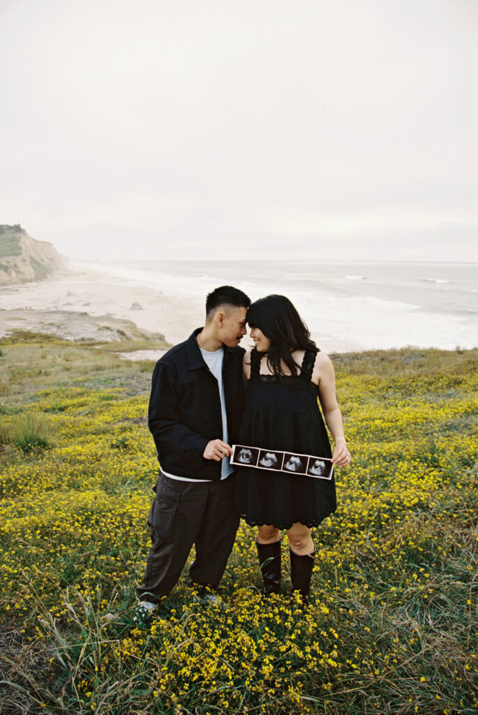 Couple holding ultrasound in yellow wildflowers at Half Moon Bay on 35mm film, Bay Area photographer Rose Freedman