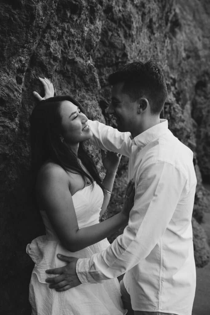 Black and white intimate couple portrait against rock wall at Rodeo Beach, Bay Area engagement photographer Rose Freedman