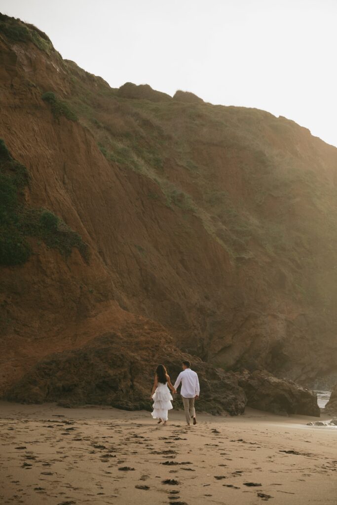 Couple walking hand in hand at Rodeo Beach with dramatic cliffs, Rodeo Beach engagement photographer Rose Freedman