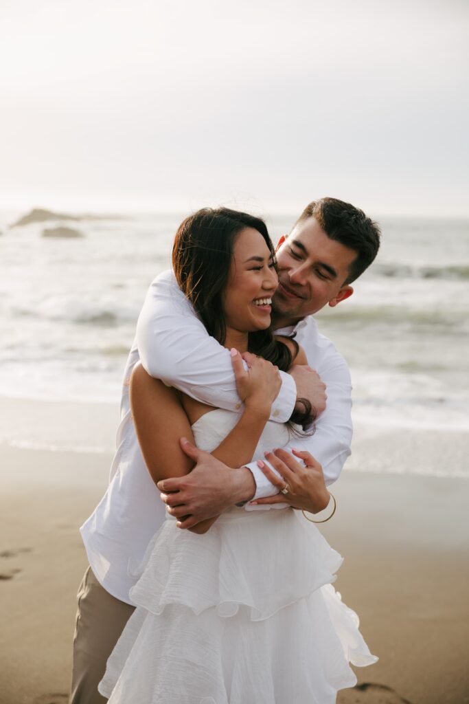 Couple laughing and embracing on Rodeo Beach at golden hour, Rodeo Beach engagement photographer Rose Freedman
