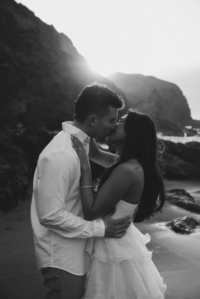 Black and white couple kissing at Rodeo Beach with dramatic cliffs and sunlight, Bay Area engagement photographer Rose Freedman
