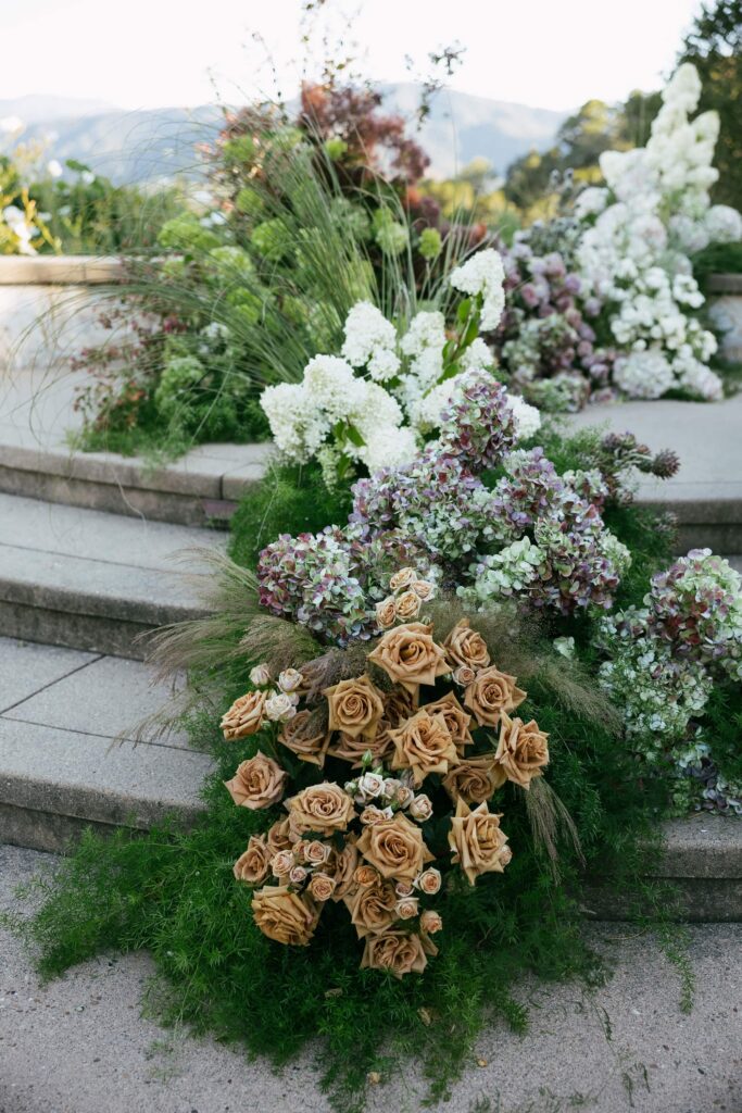 Lush floral installation cascading down stone steps at Holman Ranch ceremony — caramel roses, purple blooms, and greenery, Carmel Valley wedding photographer