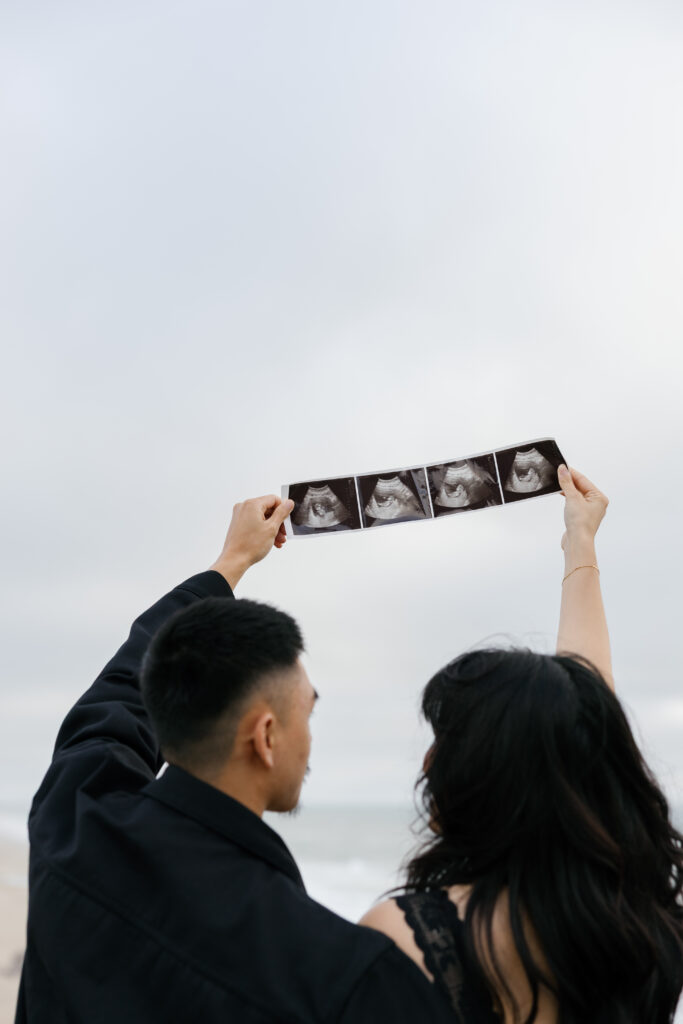 Couple holding ultrasound up to sky at Half Moon Bay coast, pregnancy announcement photographer Rose Freedman