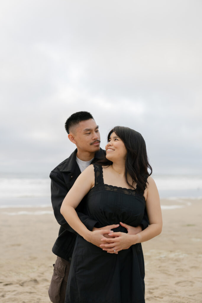 Couple embracing on Half Moon Bay beach with hands on belly, Bay Area couples photographer Rose Freedman