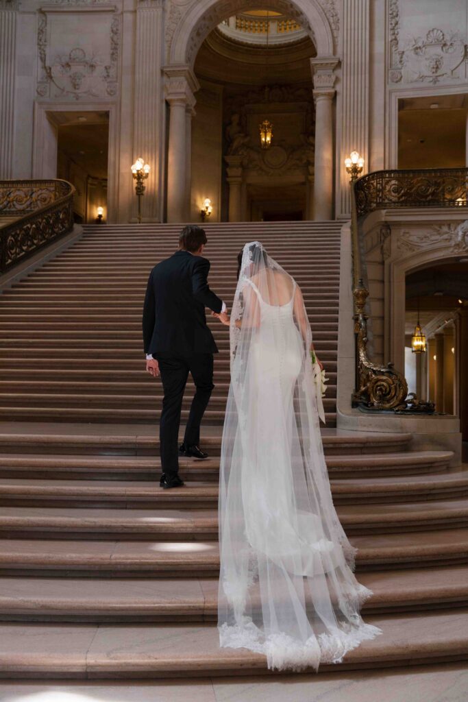 Bride with cathedral veil on grand staircase at SF City Hall, San Francisco wedding photographer Rose Freedman