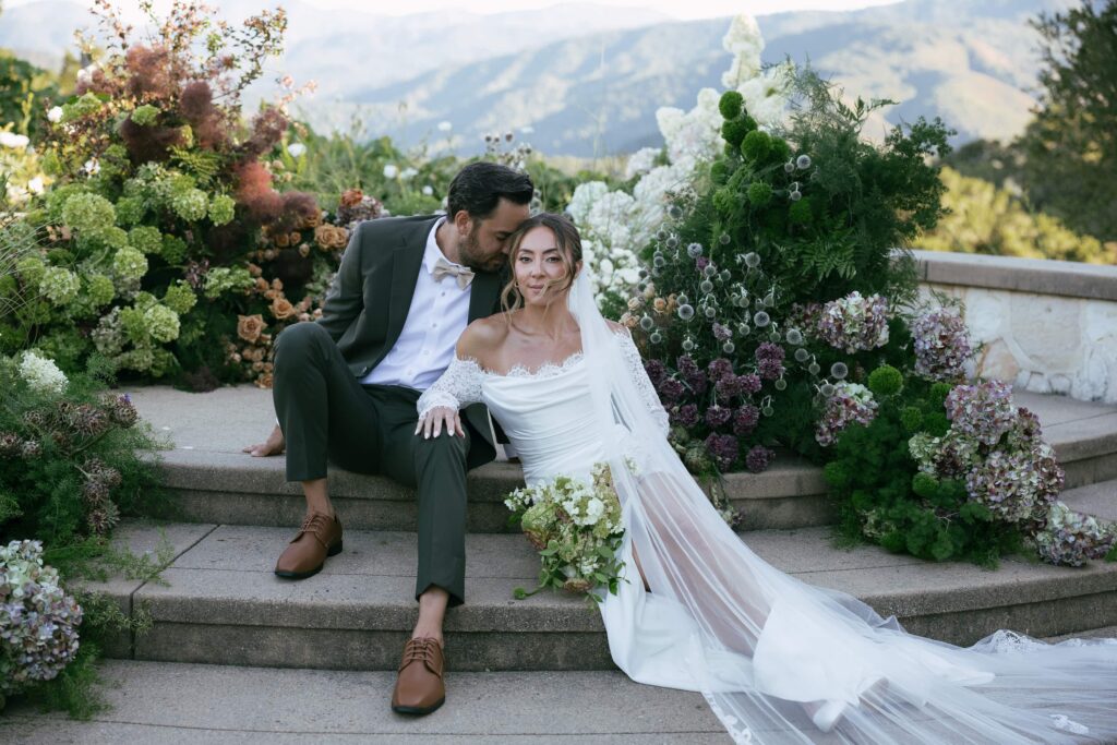 Bride and groom seated on stone steps surrounded by lush floral arrangements with Napa hills in background, Bay Area wedding photographer Rose Freedman