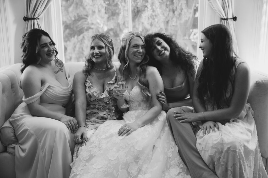 Black and white photo of bride laughing with bridesmaids on sofa getting ready, Bay Area wedding photographer Rose Freedman