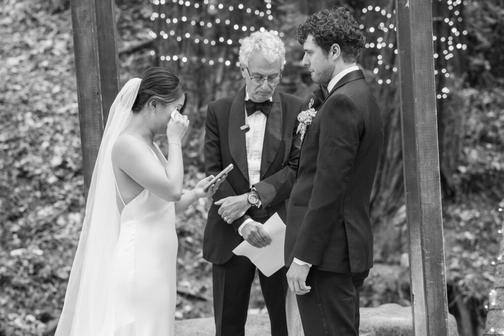 Bride wiping tears during outdoor redwood wedding ceremony at Saratoga Springs with string lights hanging between trees