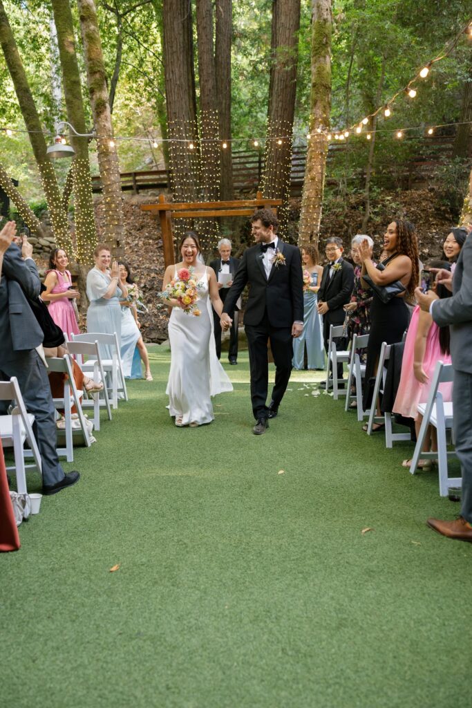Newlyweds walking back down the aisle through redwood wedding venue at Saratoga Springs as guests cheer