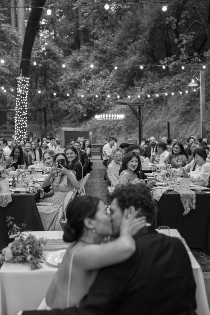 Bride and groom sharing a kiss at their sweetheart table during outdoor Saratoga Springs redwood wedding reception